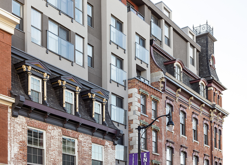 Close-up of a historic brick building with ornate detailing, including mansard roofs, dormer windows, and decorative trim. A modern addition rises above the original structure, featuring a smooth facade, large windows, and glass balconies, creating a contrast between the old and new architectural styles.