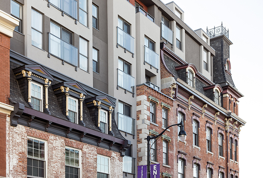 Close-up of a historic brick building with ornate detailing, including mansard roofs, dormer windows, and decorative trim. A modern addition rises above the original structure, featuring a smooth facade, large windows, and glass balconies, creating a contrast between the old and new architectural styles.