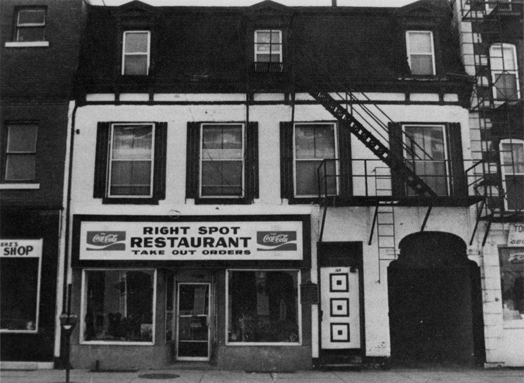 Black and white photo of a two-story building with a restaurant on the ground floor. The sign above the entrance reads ‘Right Spot Restaurant’ with a Coca-Cola logo. The facade features two large display windows and a central door. The upper floor has three windows with dark shutters, and a fire escape is visible on the right side of the building.