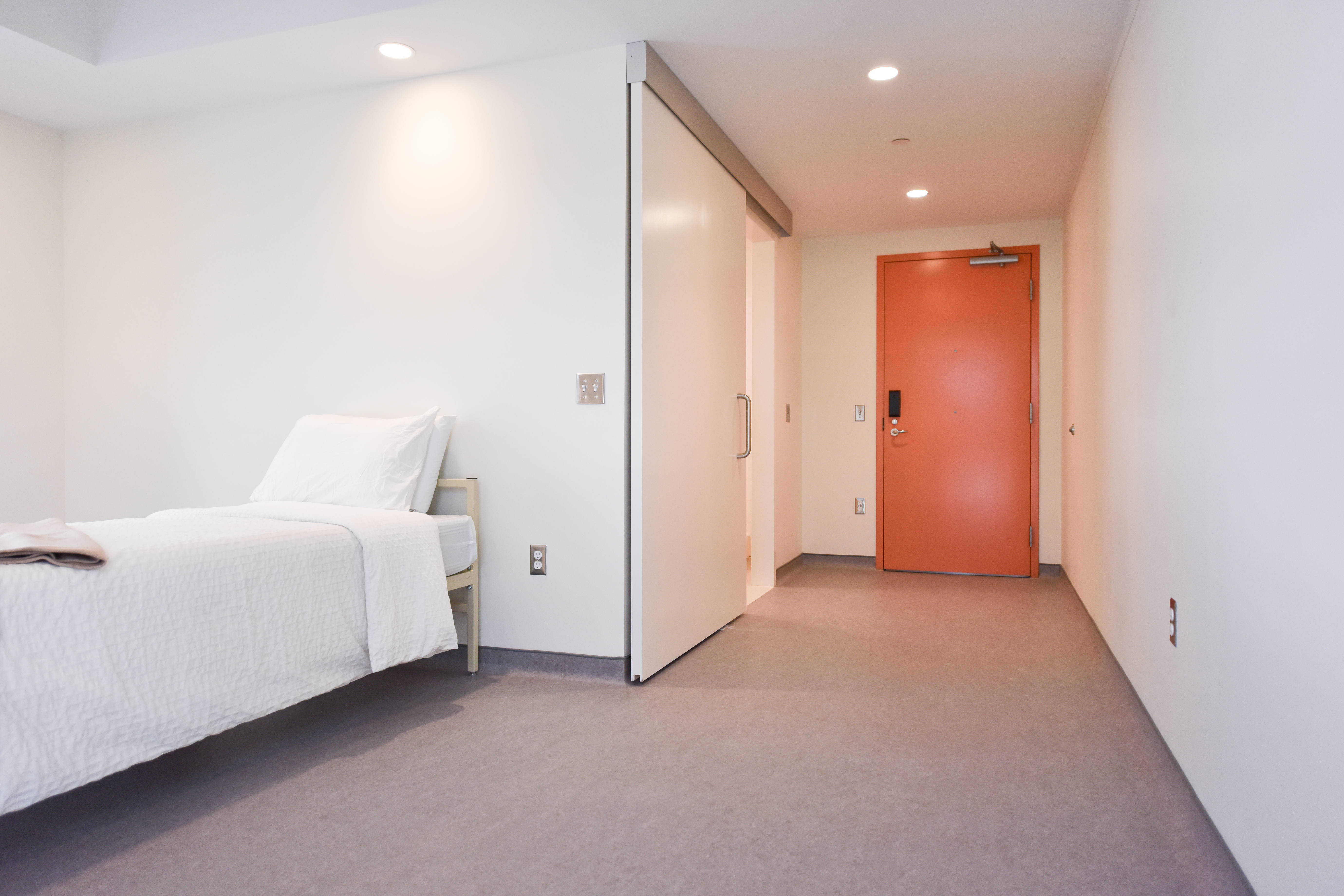 Interior of a simple bedroom featuring a single bed with white linens positioned near a white sliding door. The walls and floor are light-colored, and recessed ceiling lights provide illumination. In the background, an orange door contrasts with the otherwise neutral tones, marking the entry to the room.