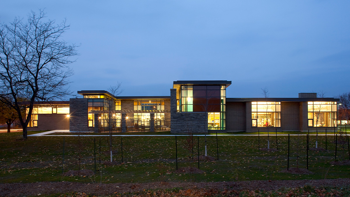 South-facing view of a modern library with warm interior lighting, large windows, and a mix of brick and stone cladding at dusk.