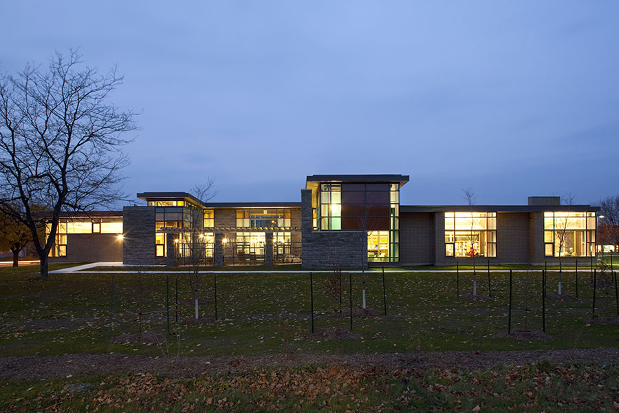 South-facing view of a modern library with warm interior lighting, large windows, and a mix of brick and stone cladding at dusk.