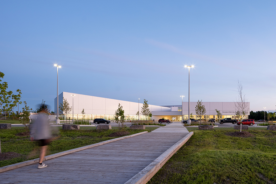 A wooden path overtop lush green grass leads to the modern looking Georgina Multi-Use Recreation Centre