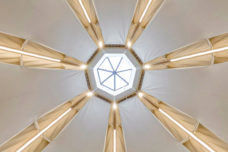 An upward view of the ceiling with a central octagonal skylight and white fabric panels, separated by illuminated wooden beams.                                                                                                                                                                                                                                                                                                                                                                                                                                                                                                                                                                                                                                                                   