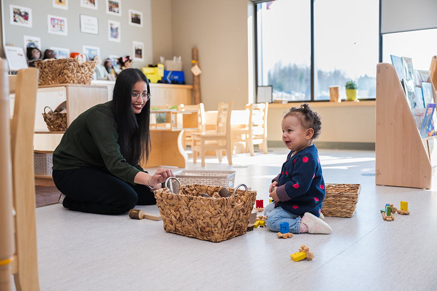 An inviting classroom where a child and adult interact on the floor with wooden blocks and baskets, surrounded by child-friendly furniture, natural light, and educational materials.