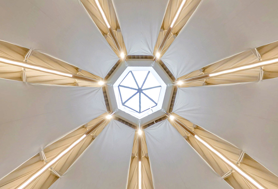 An upward view of the ceiling with a central octagonal skylight and                                                                                                                                                                                                                                                                                                                                                                                                                                                                                                                                                                                                                                                                    white fabric panels, separated by illuminated wooden beams.