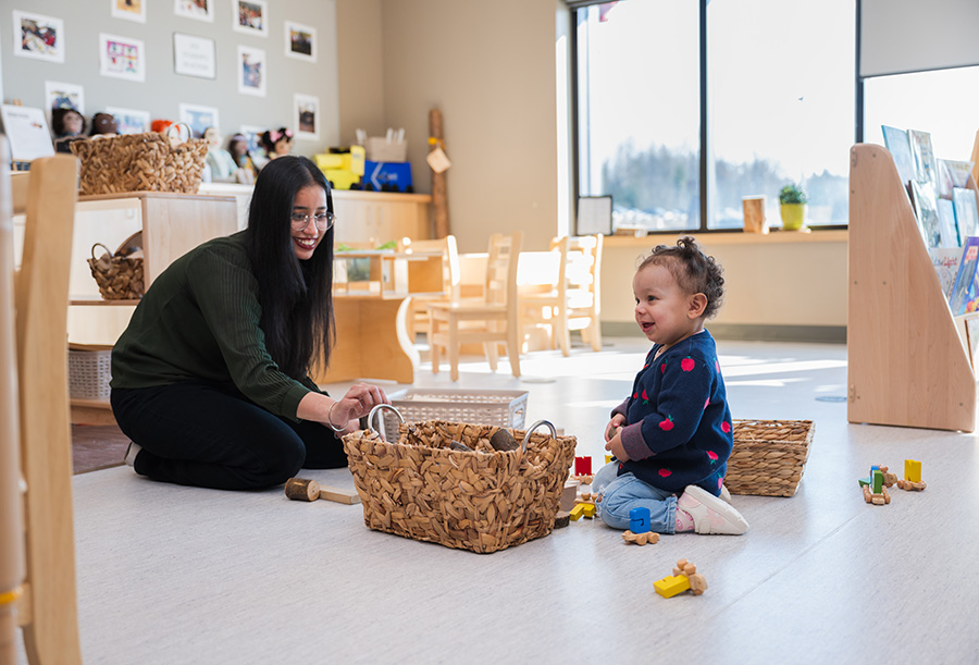 An inviting classroom where a child and adult interact on the floor with wooden blocks and baskets, surrounded by child-friendly furniture, natural light, and educational materials.